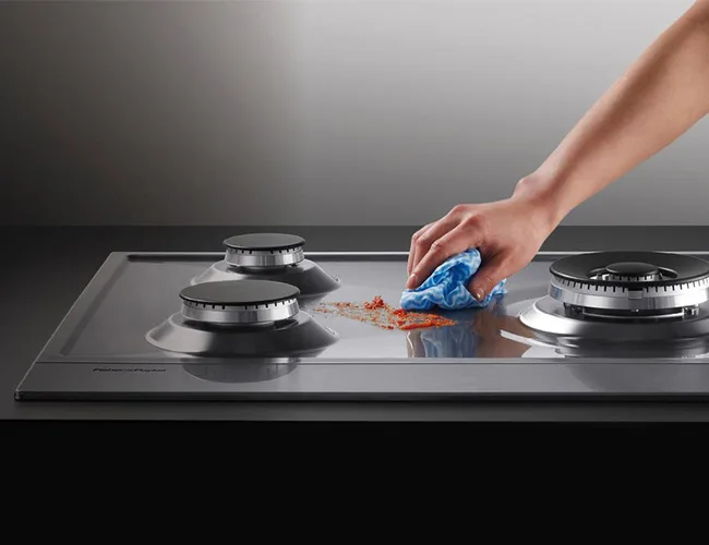 Kookplaat schoonmaken: een hand veegt met een blauw doekje een vlek weg tussen de branders van een gaskookplaat